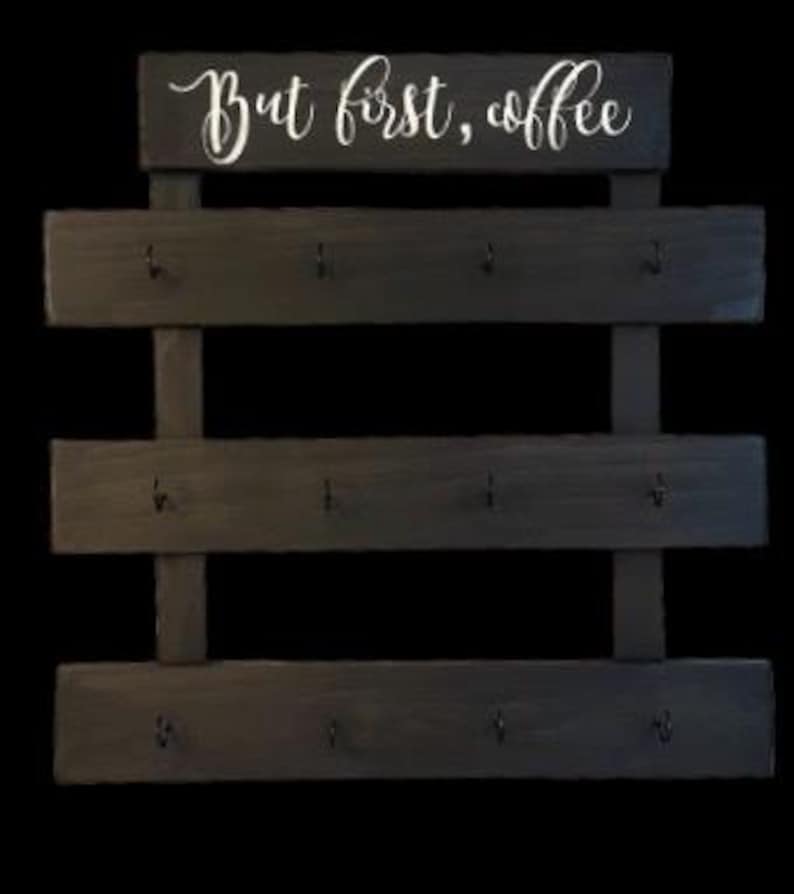 May include: A dark wood wall-mounted rack featuring three horizontal slats, each equipped with four black metal hooks. The top section displays the white cursive text "But first, coffee" printed on a rectangular wooden board.