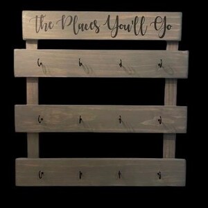 May include: A gray wooden wall-mounted organizer featuring three horizontal planks, each equipped with four black metal hooks. The top plank displays the text "the Places You'll Go" in a black cursive script font, providing a functional display.