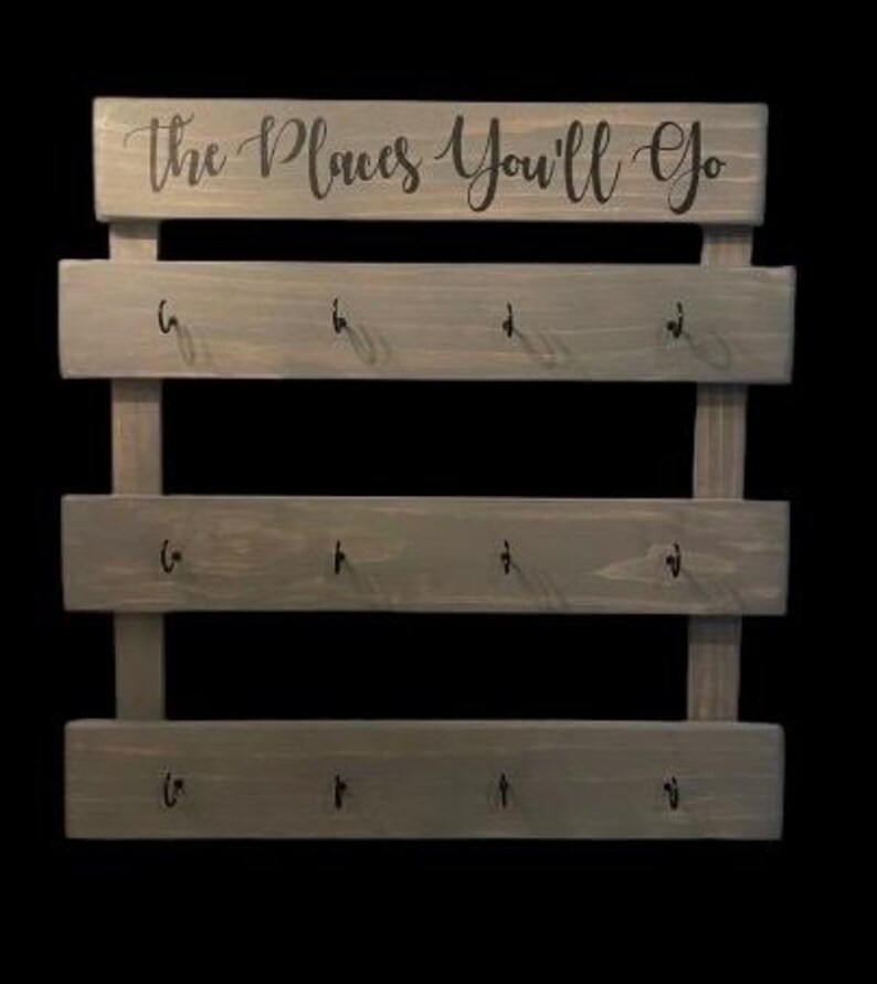 May include: A gray wooden wall-mounted organizer featuring three horizontal planks, each equipped with four black metal hooks. The top plank displays the text "the Places You'll Go" in a black cursive script font, providing a functional display.