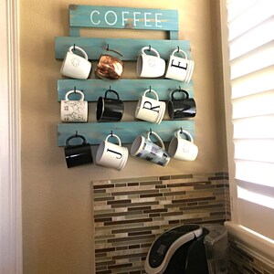 May include: A light blue wooden wall-mounted rack with the word COFFEE painted in white. The rack features three horizontal slats with black metal hooks holding eleven assorted mugs, including white, black, and metallic copper finishes.