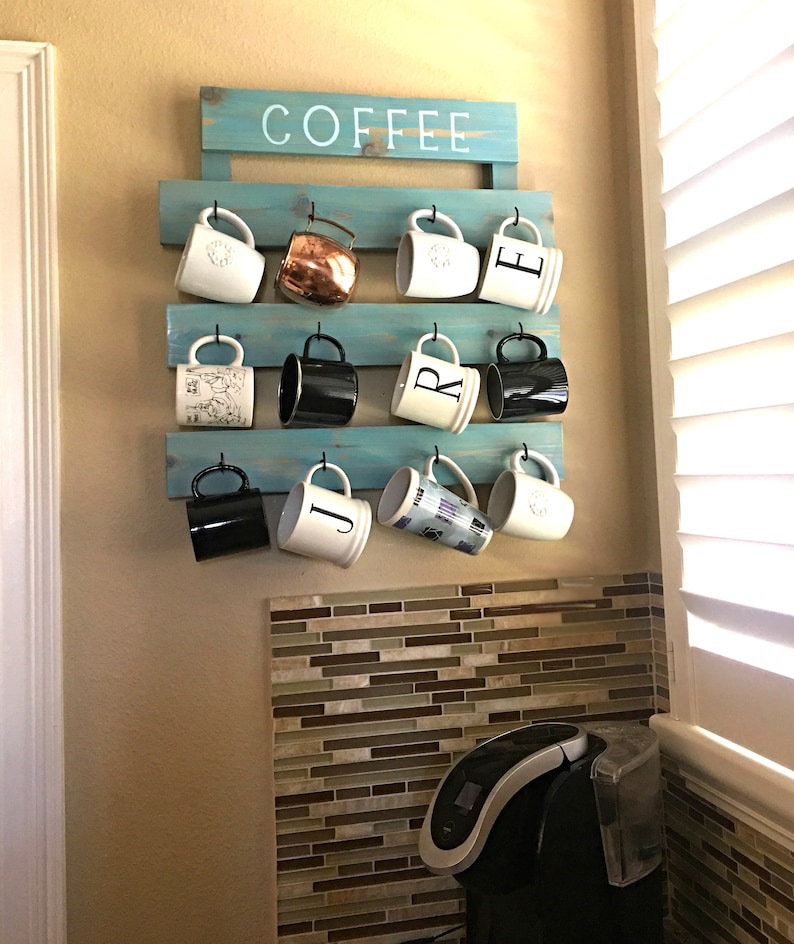 May include: A light blue wooden wall-mounted rack with the word COFFEE painted in white. The rack features three horizontal slats with black metal hooks holding eleven assorted mugs, including white, black, and metallic copper finishes.