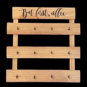 May include: A light-colored wooden wall-mounted organizer featuring three horizontal slats, each equipped with four small black metal hooks. The top slat displays the black cursive text "But first, coffee" centered across the natural wood grain surface.