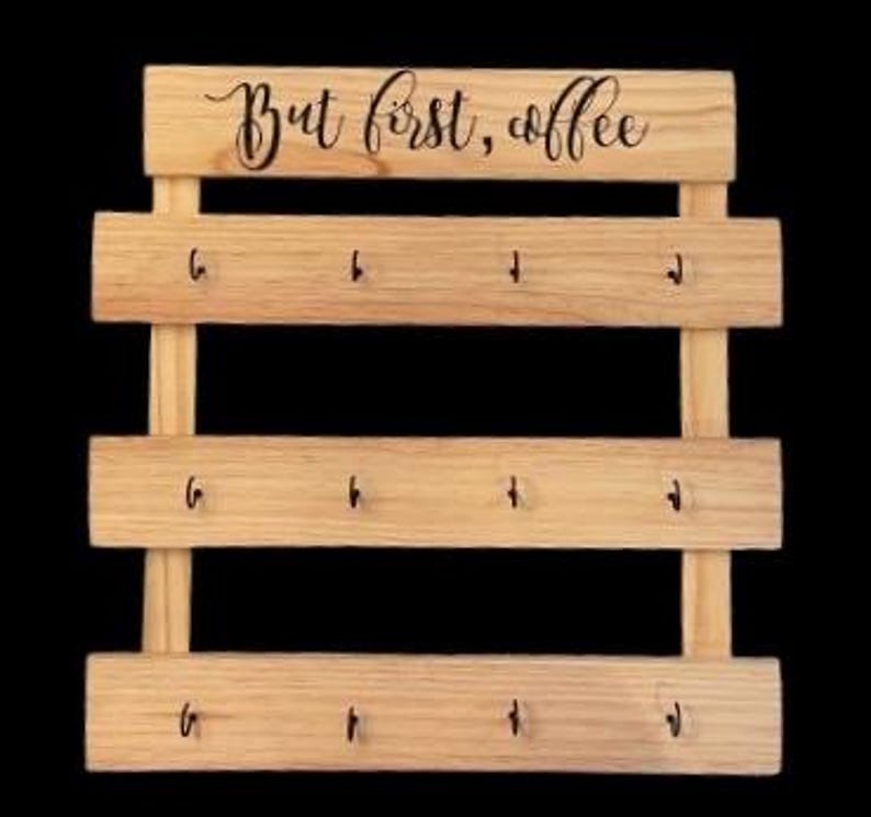 May include: A light-colored wooden wall-mounted organizer featuring three horizontal slats, each equipped with four small black metal hooks. The top slat displays the black cursive text "But first, coffee" centered across the natural wood grain surface.