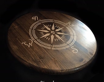 May include: A round wooden coaster with a dark brown stain. The coaster features a compass design with a white star in the center and the letters N, E, S, and W around the edge.