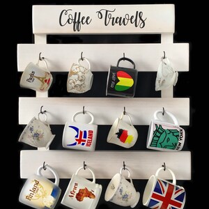 May include: A white wooden wall-mounted rack with the text Coffee Travels printed at the top. The rack features three horizontal slats with twelve black metal hooks, each holding a ceramic mug with various travel-themed designs and patterns.