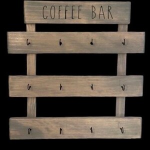 May include: A gray-stained wooden wall rack featuring the text COFFEE BAR printed in black on the top horizontal slat. The rack consists of three additional horizontal slats, each equipped with four black metal hooks for hanging items.
