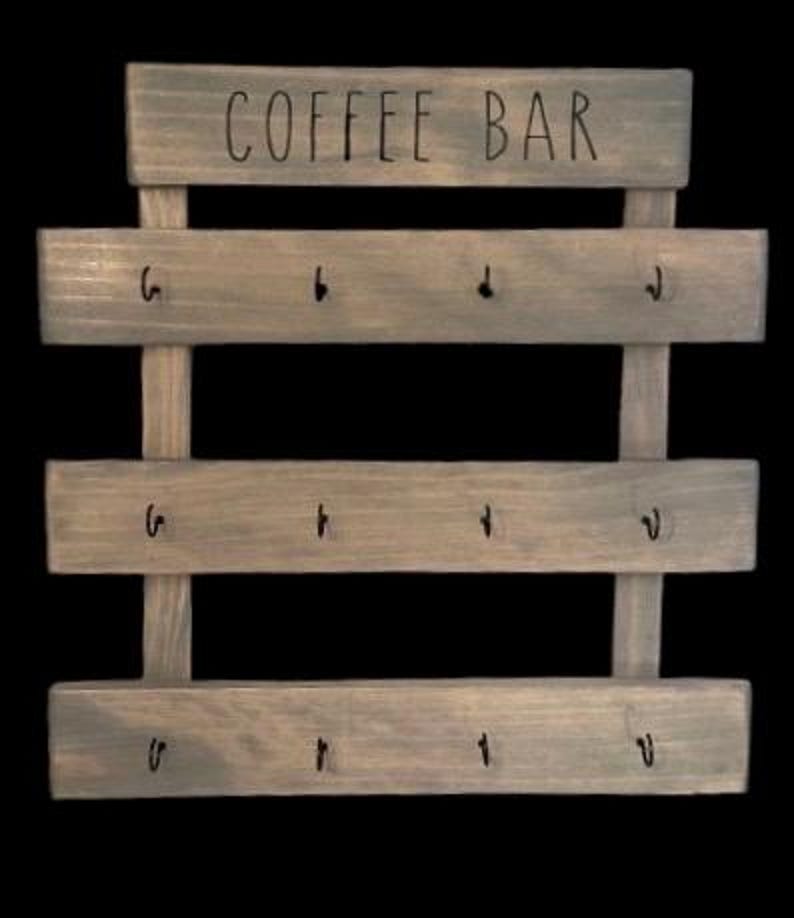 May include: A gray-stained wooden wall rack featuring the text COFFEE BAR printed in black on the top horizontal slat. The rack consists of three additional horizontal slats, each equipped with four black metal hooks for hanging items.