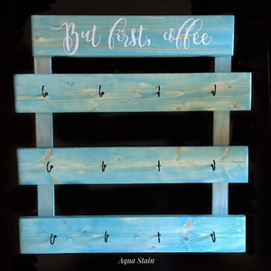 May include: A light blue stained wooden wall rack with three horizontal slats, each featuring four black metal hooks. The top slat displays the white cursive text "But first, coffee". The wood grain is visible through the aqua blue stain finish.