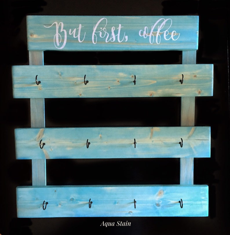 May include: A light blue stained wooden wall rack with three horizontal slats, each featuring four black metal hooks. The top slat displays the white cursive text "But first, coffee". The wood grain is visible through the aqua blue stain finish.
