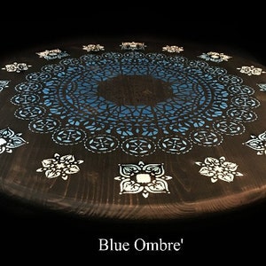 May include: A dark wood round table with a blue ombre mandala design painted on the top. The mandala has a white lace-like pattern around the edges. The table has a blue ombre effect, with the darkest blue in the center and the lightest blue at the edges.