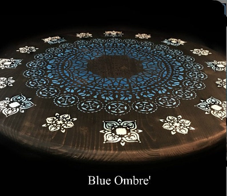 May include: A dark wood round table with a blue ombre mandala design painted on the top. The mandala has a white lace-like pattern around the edges. The table has a blue ombre effect, with the darkest blue in the center and the lightest blue at the edges.