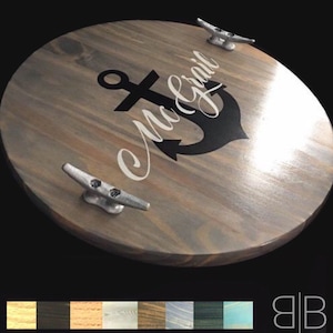 May include: A round wooden serving tray with a smooth, polished finish. The tray features a black anchor graphic overlaid with white cursive text reading McGrail. Two silver-toned metal boat cleats are attached to the top as handles.