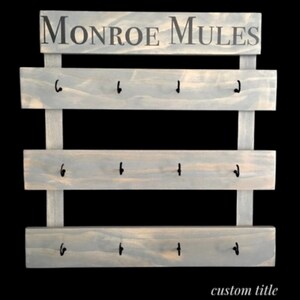 May include: A light gray wooden wall rack featuring three horizontal planks with four black metal hooks each, totaling twelve hooks. The top header board displays the text MONROE MULES in dark gray lettering. The structure is held by two vertical supports.