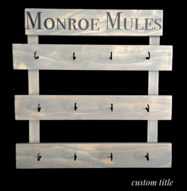 May include: A light gray wooden wall rack featuring three horizontal planks with four black metal hooks each, totaling twelve hooks. The top header board displays the text MONROE MULES in dark gray lettering. The structure is held by two vertical supports.