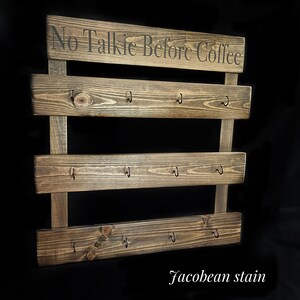 May include: A dark brown stained wooden wall rack with three horizontal planks, each featuring four black metal hooks. The top plank displays the text No Talkie Before Coffee in black lettering. The wood has a visible grain and Jacobean stain finish.