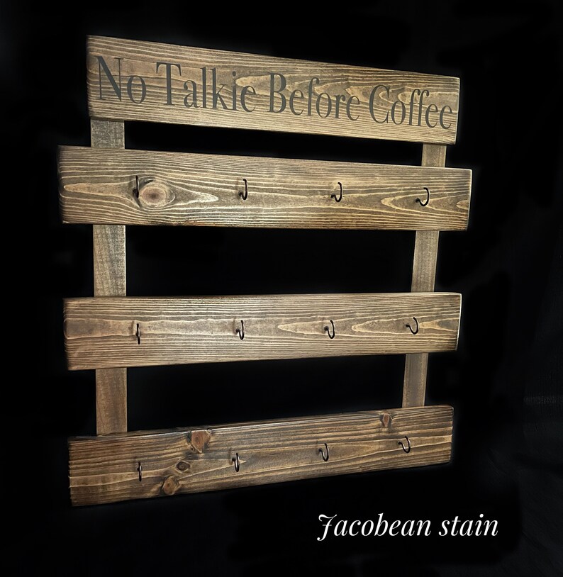May include: A dark brown stained wooden wall rack with three horizontal planks, each featuring four black metal hooks. The top plank displays the text No Talkie Before Coffee in black lettering. The wood has a visible grain and Jacobean stain finish.
