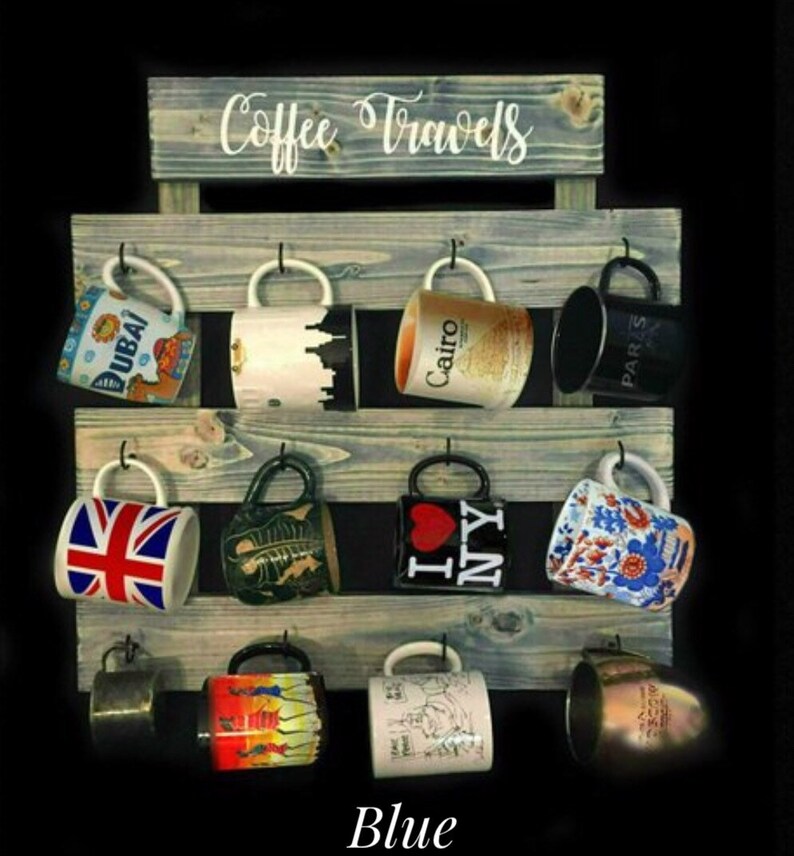 May include: A blue-toned wooden wall-mounted mug rack with the text Coffee Travels printed at the top. The rack features three horizontal slats with twelve black metal hooks, each holding a ceramic mug with various travel-themed designs.