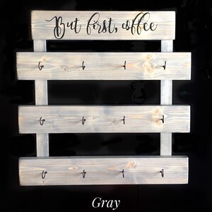 May include: A gray wooden wall-mounted rack featuring three horizontal planks with four black metal hooks each, totaling twelve hooks. The top plank displays the black cursive text "But first, coffee" and the word "Gray" is printed below.