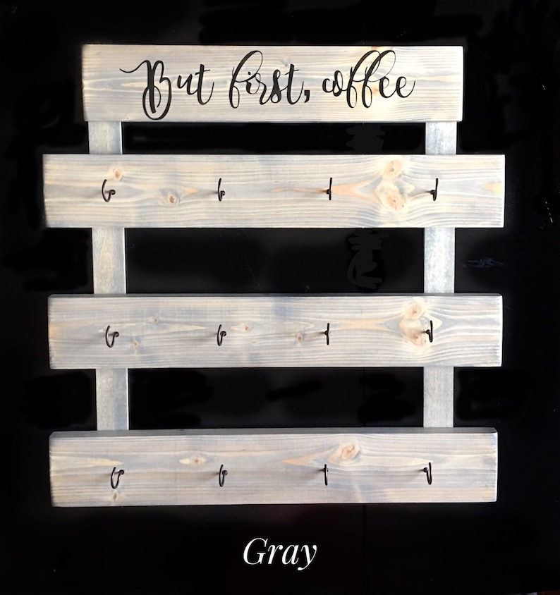May include: A gray wooden wall-mounted rack featuring three horizontal planks with four black metal hooks each, totaling twelve hooks. The top plank displays the black cursive text "But first, coffee" and the word "Gray" is printed below.