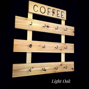 May include: A light oak wall-mounted mug rack featuring three horizontal wooden planks attached to two vertical supports. Each plank holds four black metal hooks. A rectangular wooden sign at the top displays the word COFFEE in black text.
