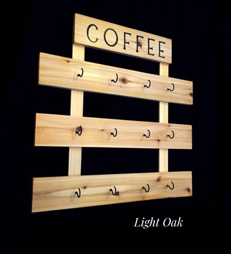 May include: A light oak wall-mounted mug rack featuring three horizontal wooden planks attached to two vertical supports. Each plank holds four black metal hooks. A rectangular wooden sign at the top displays the word COFFEE in black text.