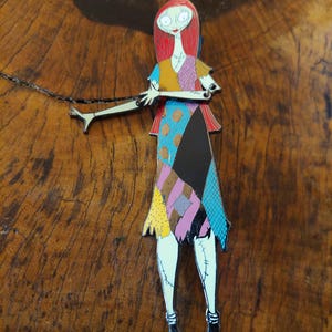 May include: A colourful enamel pin of Sally from The Nightmare Before Christmas. The pin shows Sally in her patchwork dress with red hair and pale skin. The pin is a collectible item and a fan favourite.