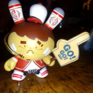 May include: A collectible figurine of a rabbit-like character in a red and white uniform, holding a foam finger sign that says "GO! GO! GO!". The figure has a brown head, yellow face, and white ears with red accents. It is standing on a wooden surface.