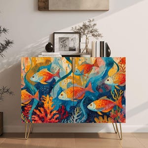 May include: A rectangular cabinet featuring a vibrant underwater scene with multiple orange and red fish swimming among colorful coral reefs. The cabinet is supported by four thin, gold-toned metal hairpin legs and has a smooth, flat front surface.