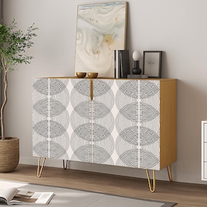 May include: A wooden cabinet with a geometric pattern on the doors. The cabinet has gold-colored hairpin legs and a light wood finish. A small tree in a woven pot and a side table with a lamp are also visible.
