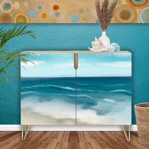 May include: A rectangular cabinet featuring a printed ocean wave design on its two doors, supported by thin gold-toned metal legs. The cabinet surface displays a blue sea with white foam and a light blue sky, creating a continuous coastal scene.