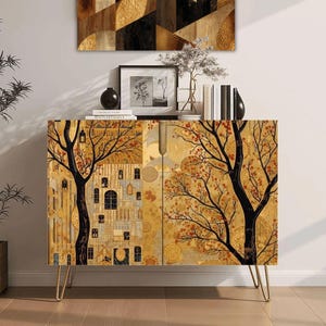 May include: A rectangular cabinet with gold, black, and brown tones featuring a detailed tree and building print on the front doors. The cabinet is supported by four thin, gold-colored metal hairpin legs and has a flat top surface.