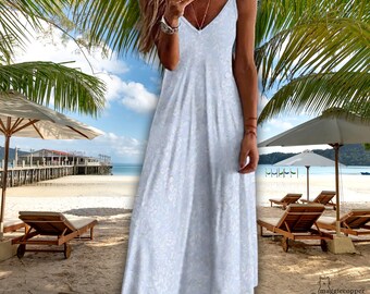 May include: A white, floral-patterned maxi dress with spaghetti straps and a V-neck. The dress is worn on a beach with palm trees, umbrellas, and lounge chairs in the background. The dress is floor-length.