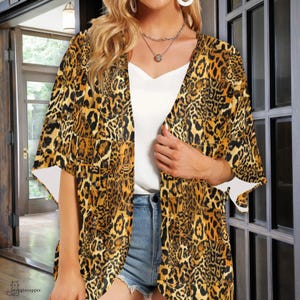 May include: A leopard print kimono-style jacket in shades of brown, black, and yellow. The open-front design features wide sleeves with white accents. The jacket is worn over a white top and denim shorts.