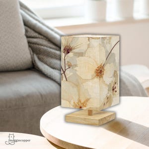 May include: A rectangular table lamp with a floral design. The lamp has a light beige lampshade with a watercolour-style flower pattern and a square wooden base. The lamp is on a white table.