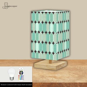 Può includere: Una lampada da tavolo rettangolare con base in legno chiaro e paralume fantasia. Il paralume presenta un motivo ripetuto di ovali turchese e grigio chiaro su sfondo marrone scuro. La lampada include un telecomando e una lampadina intelligente a LED.