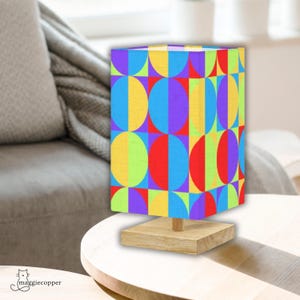 May include: A colorful table lamp with a geometric pattern. The lamp features a rectangular shade with a design of half-circles and squares in red, yellow, blue, green, and purple. It sits on a square wooden base and a wooden stem.