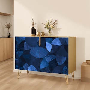 May include: A rectangular sideboard cabinet with a gold frame and four thin gold legs. The two front doors feature a dark blue and navy abstract pattern of overlapping geometric shapes. A black vase and two gold decorative bowls sit on top.
