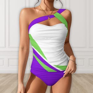 May include: A one-shoulder swimsuit in white, purple, and green. The top has a white bodice with green and purple diagonal accents. The bottom is solid purple. The swimsuit is designed with a modern, geometric pattern.