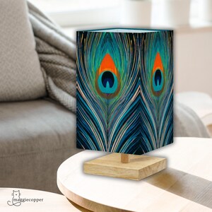 May include: A decorative table lamp featuring a peacock feather design. The lamp shade displays a vibrant pattern of blue, teal, orange, and white, set against a black background. The lamp sits on a square, light-colored wooden base.