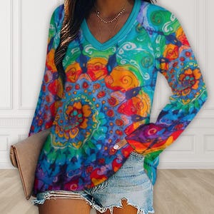 May include: A long-sleeved V-neck shirt with a vibrant, colorful tie-dye pattern. The shirt features a blue neckline and a swirling design in shades of blue, green, orange, and red. The shirt is paired with denim shorts.