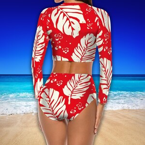 Puede incluir: Un traje de ba&ntilde;o de dos piezas con fondo rojo y estampado de hojas blancas. La parte superior de manga larga y la parte inferior de cintura alta presentan un dise&ntilde;o tropical. El traje de ba&ntilde;o se muestra sobre un fondo de playa y oc&eacute;ano.