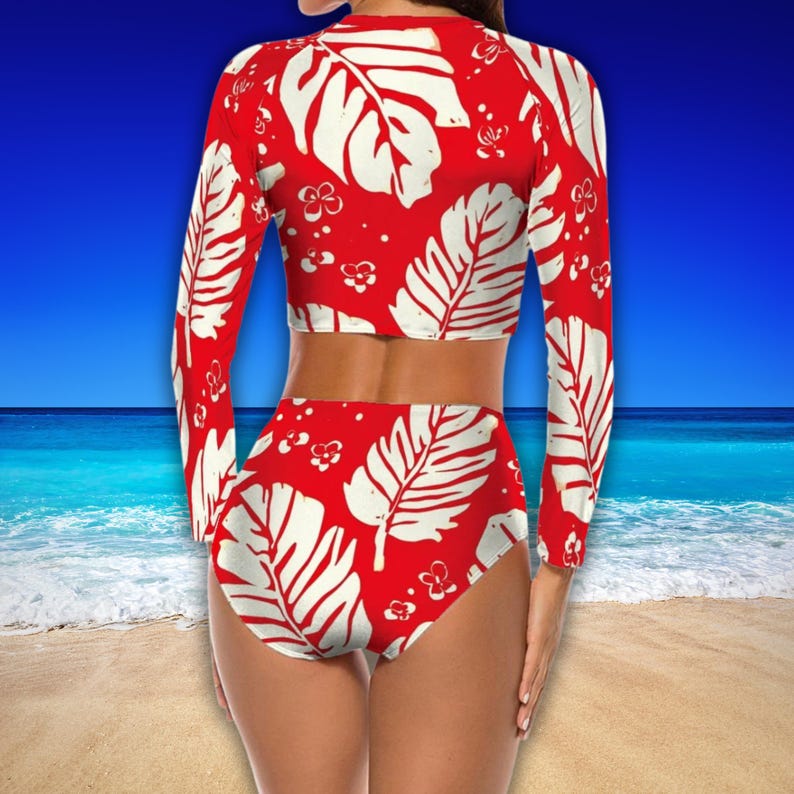Puede incluir: Un traje de ba&ntilde;o de dos piezas con fondo rojo y estampado de hojas blancas. La parte superior de manga larga y la parte inferior de cintura alta presentan un dise&ntilde;o tropical. El traje de ba&ntilde;o se muestra sobre un fondo de playa y oc&eacute;ano.