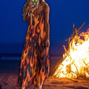 May include: A long, flowing dress with a vibrant flame pattern in shades of orange, yellow, and black. The dress has thin straps and a V-neckline. The background shows a beach scene with a bonfire.