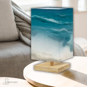 May include: A square lamp with a beach scene design. The lamp features a wooden base and a shade with a watercolor painting of ocean waves in shades of blue and white. The lamp is on a wooden table.