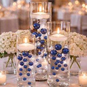 May include: Three clear cylindrical glass vases filled with water, floating white candles, and submerged glossy royal blue spheres of varying sizes. Two smaller clear glass vases contain white hydrangea flowers, and two small votive candles.