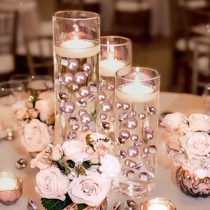 Peut inclure: Trois cylindres en verre transparent avec des bougies flottantes, des perles roses et des cristaux. Les cylindres sont entourés de roses roses et de petits bougeoirs, créant un centre de table élégant pour un mariage ou un événement spécial.
