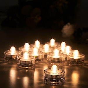 May include: A group of 12 clear plastic battery-operated LED tea lights. The lights are arranged in a semi-circle on a dark surface. The lights are all on and emitting a warm white glow.