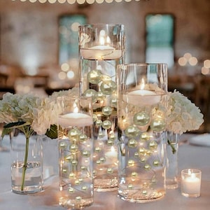 May include: Centerpiece arrangement featuring clear glass cylinders with floating candles, pearl-like beads, and fairy lights. White hydrangea flowers are placed around the cylinders. A small candle in a glass holder is also present.