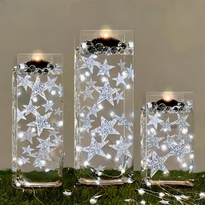 May include: Three clear glass candle holders of different heights, adorned with silver glitter stars and fairy lights. Each holder features a candle at the top, encircled by decorative beads. The holders are placed on a green surface.