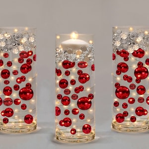 May include: Three clear glass cylinder vases decorated with red baubles, clear crystals, and fairy lights. One vase has a lit white candle. The vases are arranged on a light grey surface, creating a festive holiday display.